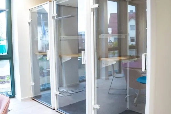 Glasfassade mit Eingangstüren eines Büros, im Hintergrund ein Tisch mit Stuhl in einem Besprechungsraum