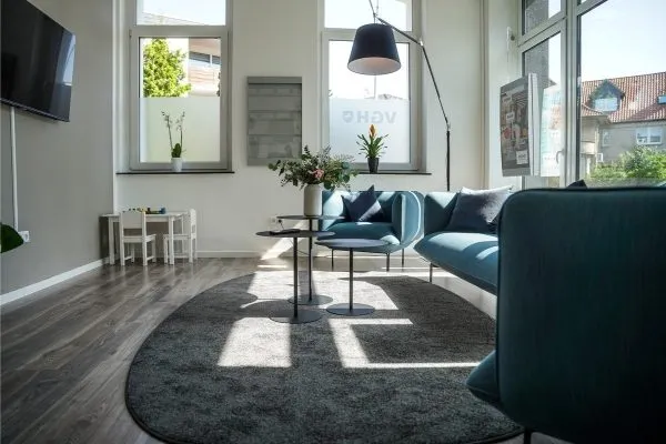 Wohnlicher Loungebereich mit petrolfarbenem Sofa, runden Tischen und Stehlampe vor Fenstern